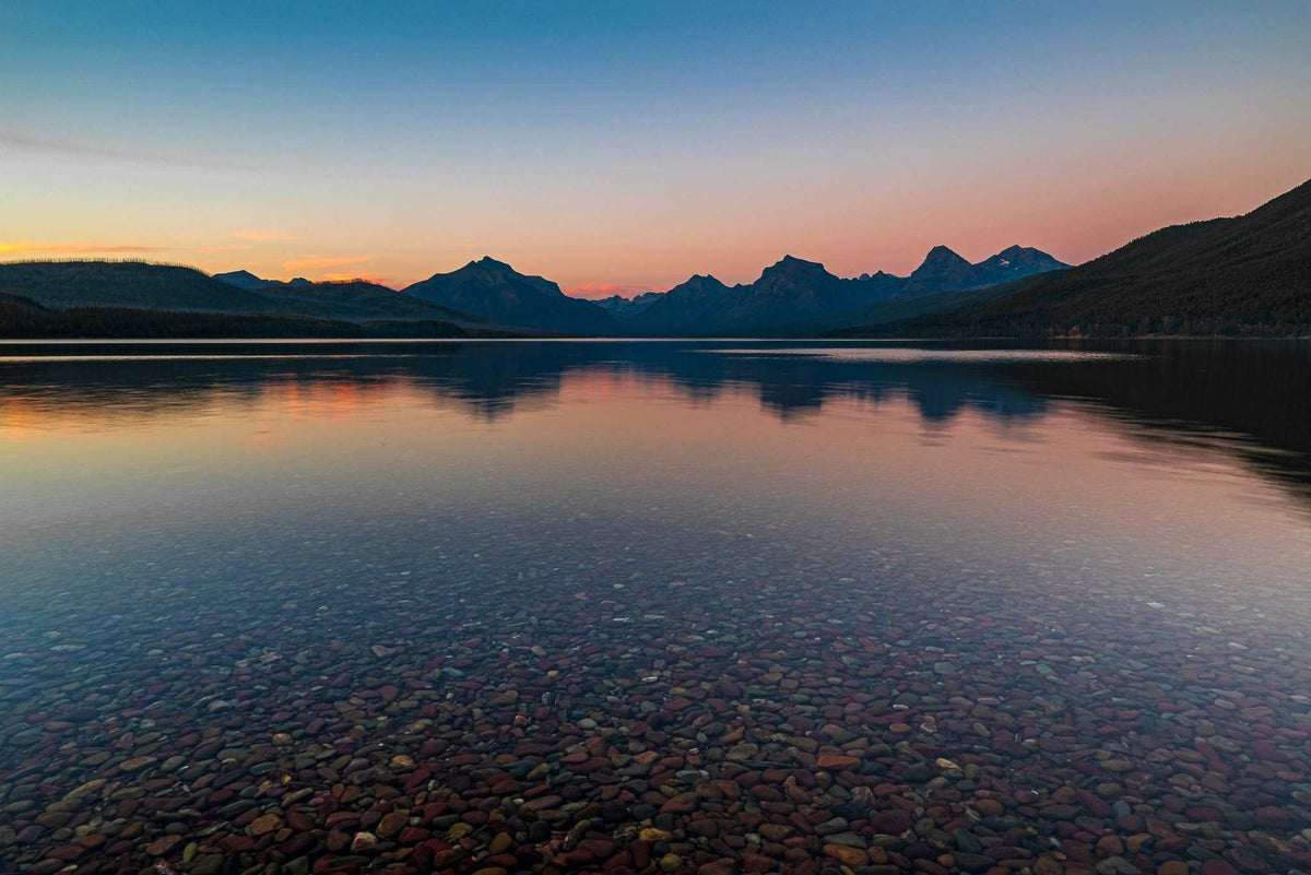 Glacier National Park Print showing a calm mountain lake at sunset, with pink-orange reflections over clear water and colorful stones