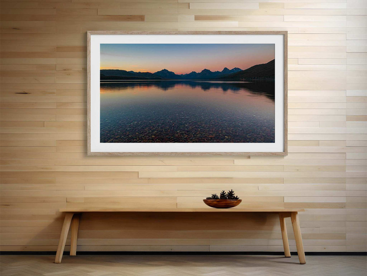 Glacier National Park Print shows a calm lake at sunset with dark mountain silhouettes, pink-orange sky, and rocky shallows