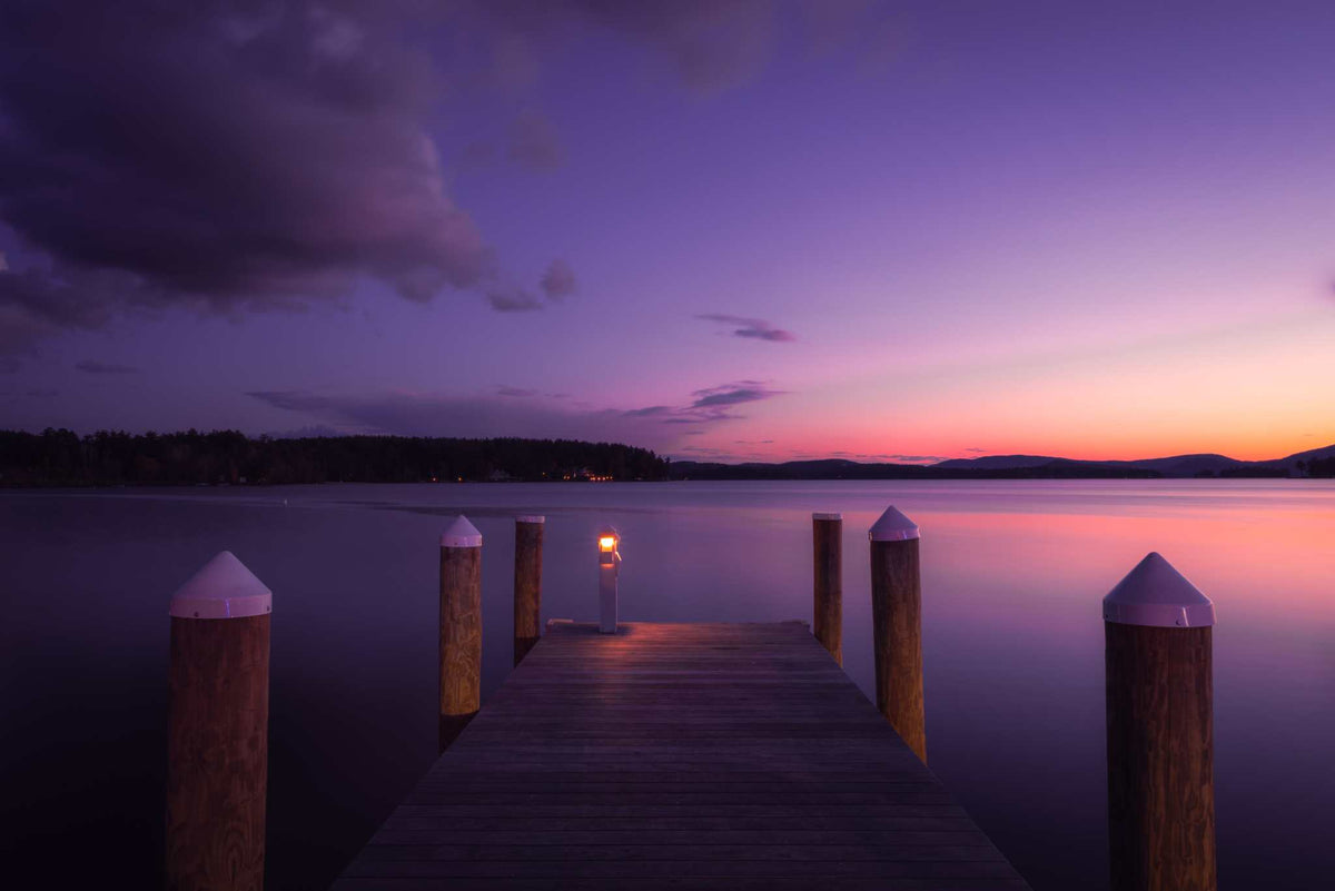 Lake Winnipesaukee Sunset Photography print with a wooden dock and glowing lamp beneath a purple, pink and orange sky