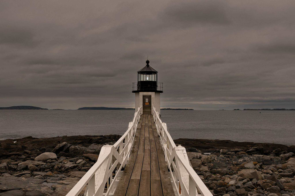 Marshall Point Lighthouse Print showing a white wooden walkway leading to a black-and-white lighthouse over a rocky coast beneath storm clouds