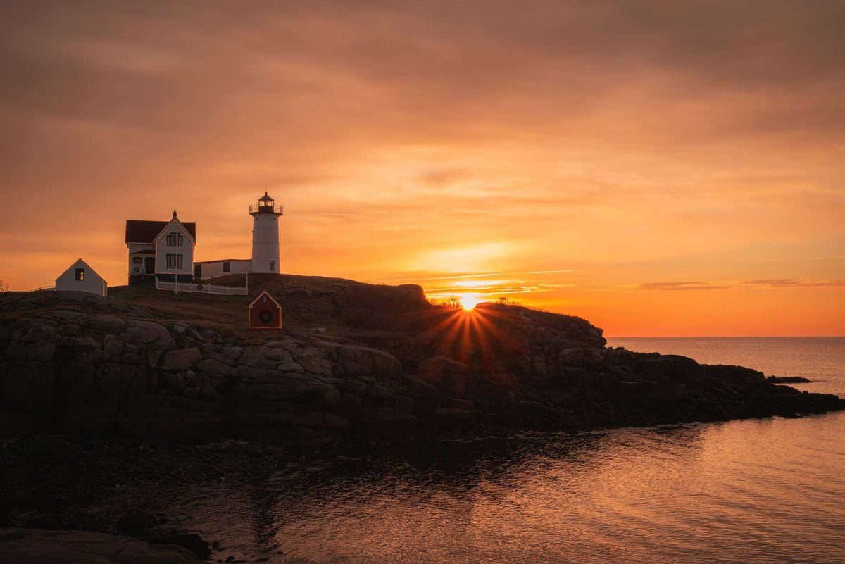 Nubble Lighthouse Print showing a white lighthouse and buildings on a rocky coast beneath a vivid orange sunrise