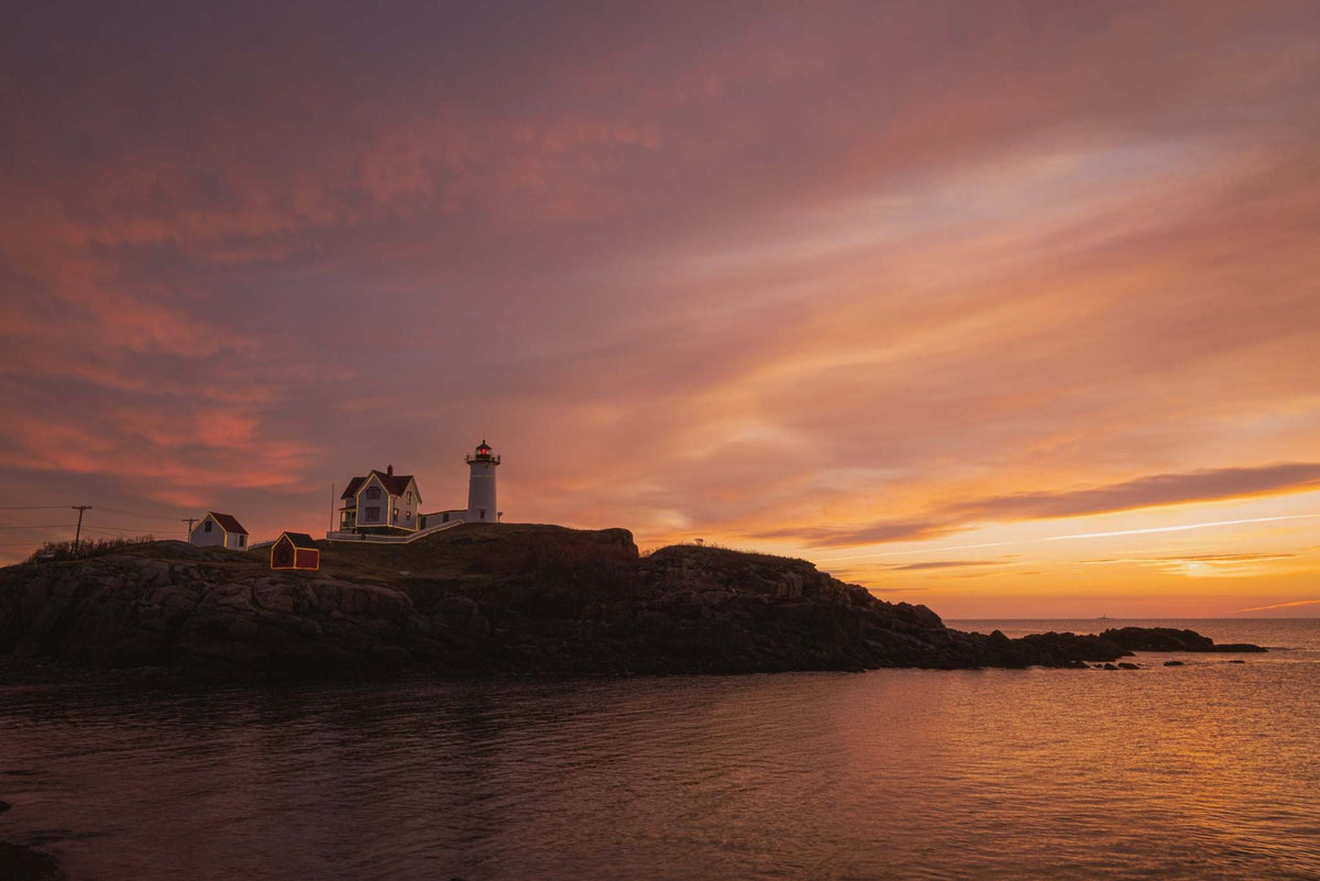 Nubble Lighthouse Wall Art showing a white lighthouse and coastal buildings on rocky shore beneath a vivid orange-pink sunrise